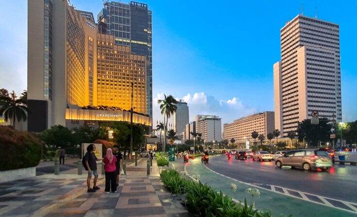 Urban street scene outside Grand Hyatt hotel with pedestrians, palm trees, and traffic at dusk/dawn.