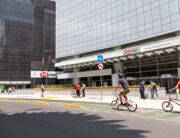 Modern city scene with a glass office building, HSBC signage, and people riding red bikes along a bike lane by the sidewalk of a busy street.