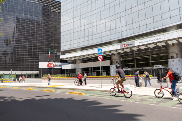 Modern city scene with a glass office building, HSBC signage, and people riding red bikes along a bike lane by the sidewalk of a busy street.