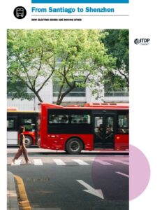 Red city bus driving past a pedestrian crossing with a woman walking on the crosswalk, trees and a modern building in the background; urban transit scene.