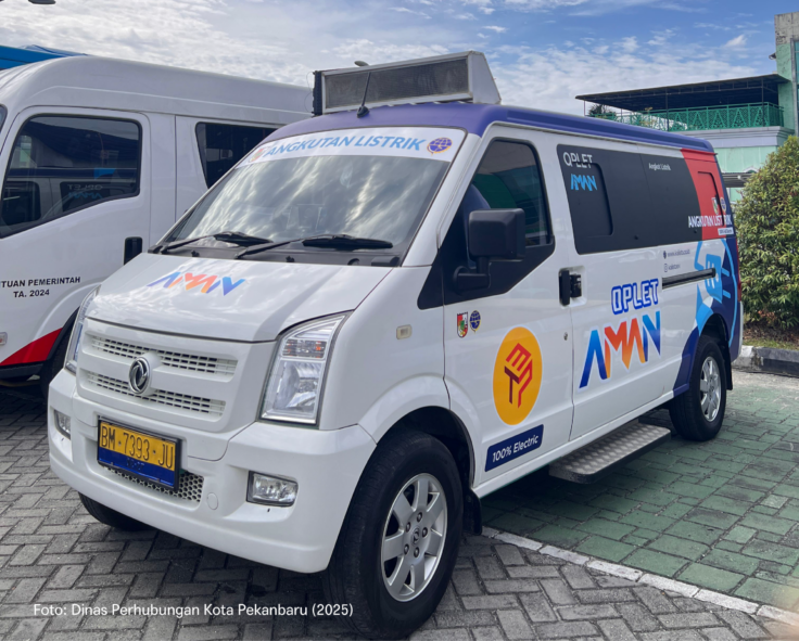 White electric van with blue accents and 'Angkutan Listrik' branding parked beside another white van; license plate BM 7393 JU visible.